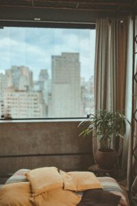 A stylish urban bedroom with cozy pillows and a stunning cityscape view through a large window.