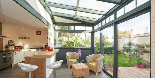 Véranda avec baies vitrées aluminium, coin salon et cuisine ouverte donnant sur le jardin.
